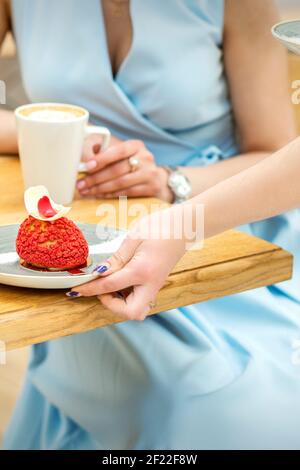 Die Hand des Kellners legt einen Teller mit auf den Tisch Ein roter Cupcake auf dem Hintergrund einer Kundin In einem Café Stockfoto