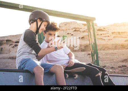 Zwei kleine Jungen mit Smartphone im Skatepark, sitzen auf einer Halfpipe Rampe. Jugendliche kommunizieren mit Freunden mit Handy-Sharing-Video von Stockfoto