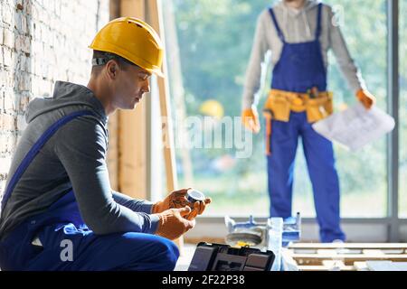 Junger Ingenieur mit Overalls und Hartmütze, der auf der Baustelle des Landhauses arbeitet und an einem sonnigen Tag Klebeband aus der Werkzeugkiste mit verschwommenem Kollegen im Hintergrund pflückt. Gebäudekonzept Stockfoto