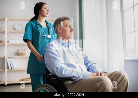 Trauriger älterer Mann im Rollstuhl, der sich deprimiert fühlte, aus dem Fenster schaute, junge Krankenschwester, die ihm zu Hause half Stockfoto