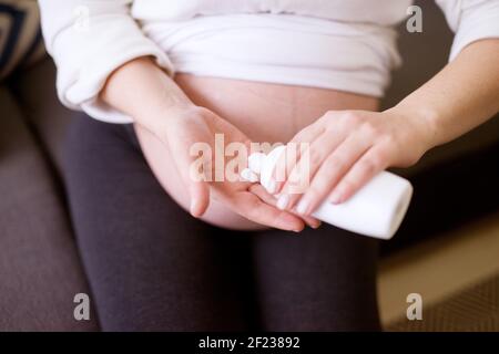 Nahaufnahme Fokus Ansicht der schwangeren Frau Hände Vorbereitung einer Creme für die Anwendung auf ihrem Bauch, während sie zu Hause sitzen. Stockfoto