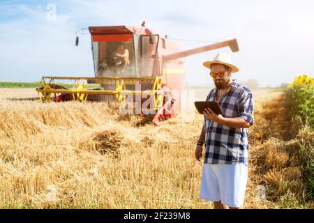 Foto eines modernen Bauernmanns mit Sonnenbrille und Hut, der den Fortschritt auf seinem Tablet überprüft, während er vor einem roten Mähdrescher steht. Stockfoto