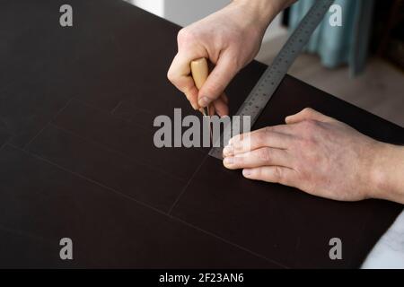 Herrenhand, die eine Ahle und ein Metalllineal hält und an einer Ledertasche in seiner Werkstatt arbeitet. Arbeitsprozess mit einem braunen Naturleder. Handwerker Stockfoto