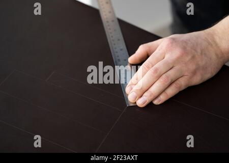 Herrenhand, die eine Ahle und ein Metalllineal hält und an einer Ledertasche in seiner Werkstatt arbeitet. Arbeitsprozess mit einem braunen Naturleder. Handwerker Stockfoto