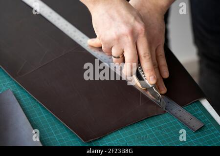 Herrenhand hält ein Schreibwarenmesser und ein Metalllineal und schneidet auf einem Stück für eine Ledertasche in seiner Werkstatt. Arbeitsprozess mit einem braunen Stockfoto