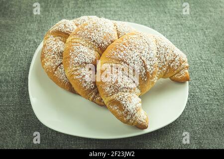 Drei Croissants auf einem weißen Teller zum Frühstück. Frische Croissants mit Puderzucker Stockfoto