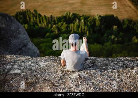 Rückansicht eines jungen Mannes, der Tee oder Kaffee in den Bergen trinkt. Reisender Mann mit Bart hält in den Händen eine Tasse Tee, nach dem Wandern. Reisen, Lifestyle, VA Stockfoto