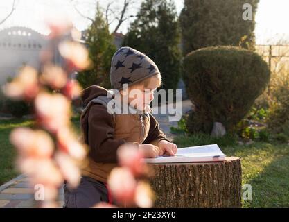 Netter Junge 4-5 Jahre alt im Hof liest oder untersucht ein Buch. Schöne, sonnige Hintergrundbeleuchtung. Fernunterricht während der Quarantänezeit, Heimunterricht Stockfoto