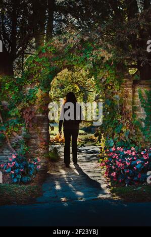 Junge Frau, die durch einen Bogen in einer Steinmauer geht In einem Garten Stockfoto