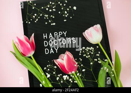 Schwarzer Briefbogen mit weißen Plastikbuchstaben mit Zitat Happy Doctor's day und einem Bündel rosa Tulpen auf rosa Hintergrund. National Doctors Day. Glücklicher Krankenschwester Tag. Flach lag Stockfoto
