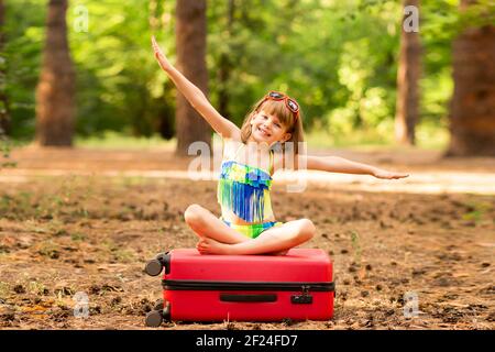 Kleines Kind Mädchen tragen Badeanzug sitzen auf Koffer im Sommerwald mit Händen nach oben, zeigen Flugzeug. Traumreise. Urlaubskonzept Stockfoto