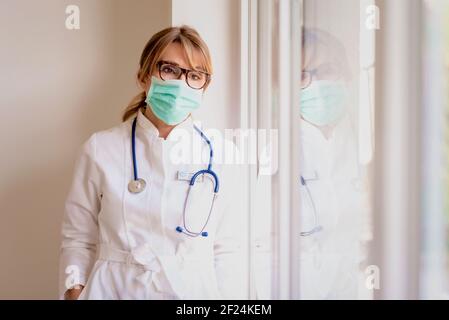 Ärztin trägt Gesichtsmaske und sieht aus gehemmten, während sie am Fenster steht. Stockfoto