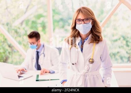 Ärztin trägt Gesichtsmasken zur Vorbeugung, während sie im Büro des Arztes steht. Männlicher Arzt arbeitet im Hintergrund. Stockfoto