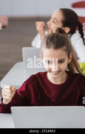 Aufgeregt schulmädchen zeigt gewinnen Geste in der Nähe Laptop im Klassenzimmer Stockfoto