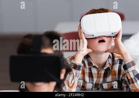 Überrascht Junge mit vr Headset in der Nähe Klassenkamerad auf verschwommenem Vordergrund Stockfoto