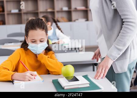 Schüler schreiben im Notebook in der Nähe von Apfel, Smartphone und Lehrer im Klassenzimmer Stockfoto