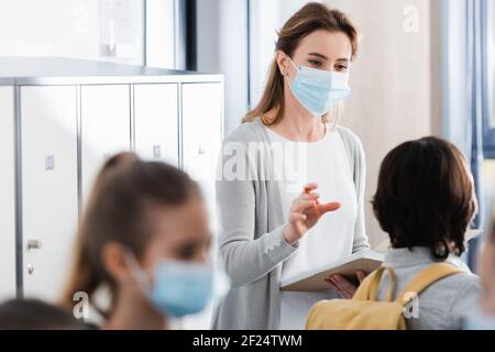 Lehrer in der medizinischen Maske hält Notizbuch und Blick auf Schüler Bei unscharfem Vordergrund Stockfoto