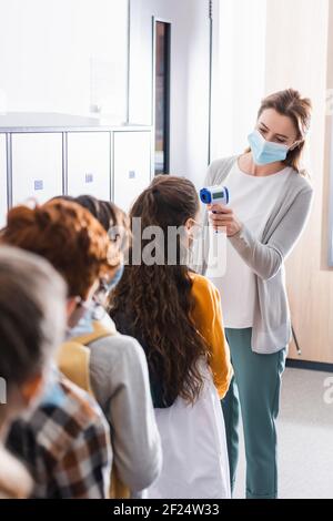 Lehrer mit berührungslosen Thermometer auf Schüler in medizinischen Masken In der Schule Stockfoto
