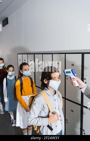 Lehrer mit Infrarot-Thermometer auf Schuljunge in Schutzmaske in der Nähe Kinder auf unscharfem Hintergrund Stockfoto