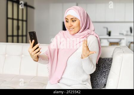 Junge muslimische arabische Mischlingsfrau, die online auf dem Handy spricht, auf der Couch im Studio sitzt, mit Familienmitarbeitern Videoanrufe macht, chattet, gestikelt, mit einem Freund über Neuigkeiten diskutiert Stockfoto