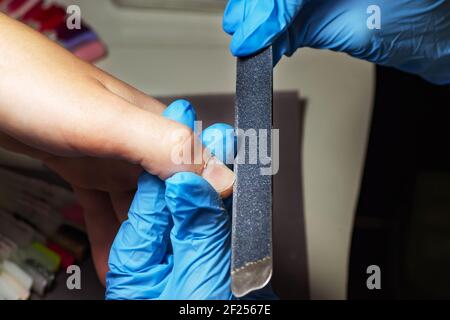 Blue-gloved Manikurist feilen einen Nagel mit einer Einweg-Feile zu Formen. Professionelle Maniküre in einem Schönheitssalon. Handpflegekonzept Stockfoto