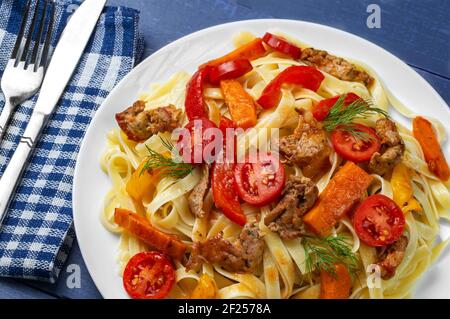 Fettuccine mit gegrilltem Fleisch, Karotten, Paprika, Dilltomaten auf blauem Holzhintergrund. Pasta mit Gemüse. Blick von oben. Stockfoto