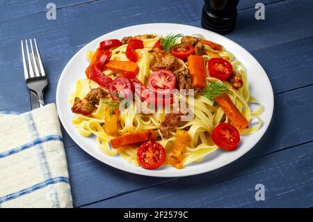 Pasta mit Gemüse. Fettuccine mit gegrilltem Fleisch, Karotten, Paprika, Dilltomaten auf blauem Holzhintergrund. Seitenansicht. Stockfoto