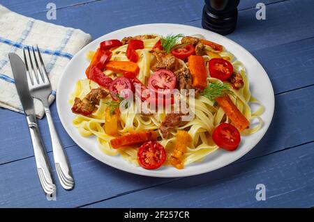 Fettuccine mit gegrilltem Fleisch, Karotten, Paprika, Dilltomaten auf blauem Holzhintergrund. Pasta mit Gemüse. Seitenansicht. Stockfoto