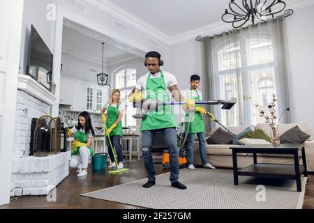Junger, hübscher, verrückter, schwarzhäutiger Putzmann in Uniform, der Spaß hat, Kopfhörer zu tragen, Musik zu hören und mit Staubsauger zu tanzen Stockfoto