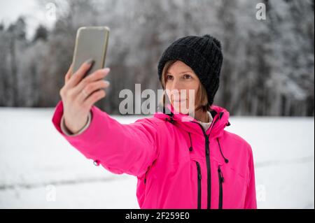 Junge Frau in rosa Winterjacke und schwarzem Hut, die ein Selfie mit ihrem Telefon in der schönen verschneiten Natur stehen. Stockfoto