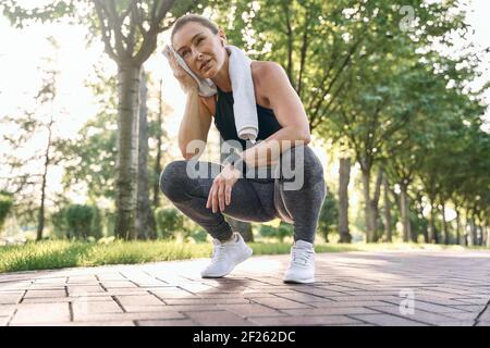 Hör nicht auf. Müde sportliche Frau mittleren Alters in Sportswear wischte ihren Schweiß mit einem Handtuch nach dem Laufen in einem grünen Park an einem sonnigen Tag Stockfoto