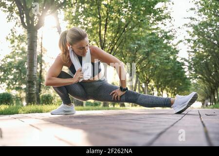 Sei fit. Attraktive sportliche Frau mittleren Alters in Sportkleidung mit Handtuch um den Hals Strecken ihre Beine während des Trainings in einem grünen Park auf einem sonnigen Stockfoto