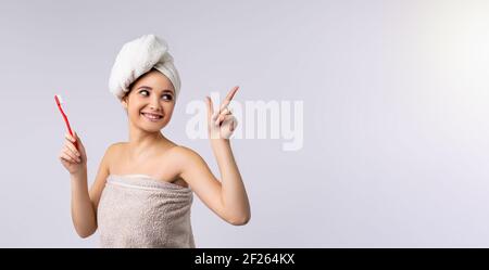 Banner, Langformat auf grauem Hintergrund. Porträt einer lächelnden hübschen Frau, die ein Badetuch trägt, die Zahnbürste hält, wegschaut und den Zeigefinger zeigt Stockfoto