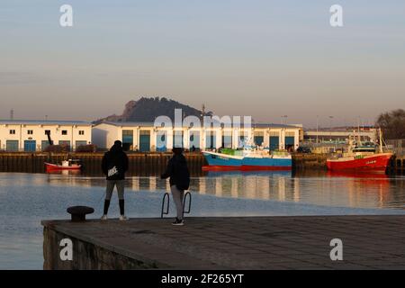 Zwei junge Männer fischen im Maliano Dock mit dem französischen Trawler Cap Finistere und dem spanischen Trawler Sukari Primero, der in der Wintermorgendssonne vor Anker liegt Stockfoto