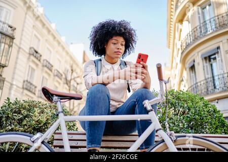 Latina Frau mit afro Frisur sitzt auf einer Bank und schaut auf ihr Handy. Hochwertige Fotos Stockfoto