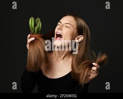 Frustrierte Frau mit langen seidig geraden Haaren in schwarzem Körper Schreiend schreiend hält Kaktuspflanze Vergleich mit Spliss Stockfoto