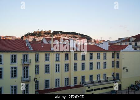 Blick auf Lissabon Sao Jorge Schloss vom Rossio Bahnhof mit gelbem Gebäude im Vordergrund bei Sonnenuntergang, in Portugal Stockfoto