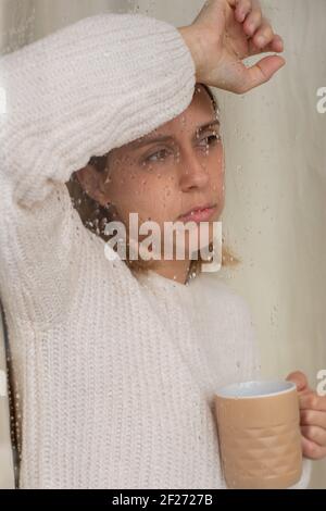 Die junge kaukasische Frau, mit kalifornischen Glanzlichtern im Haar, blickt mit nostalgischer Geste durch das nasse Fenster ihres Wohnzimmers auf den Regen. Stockfoto