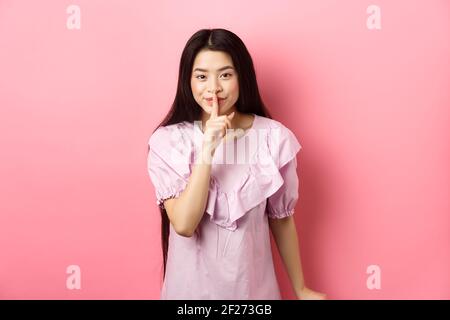 Nettes asiatisches Mädchen versteckt Geheimnis, mit Finger auf die Lippen gedrückt und lächelnd, bitten, ruhig zu bleiben, im Kleid auf rosa stehen Stockfoto