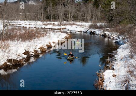 Zwei reife Frauen paddeln Kajaks auf dem Kunjamuk River in der Nähe von Speculator, NY USA in den Adirondack Mountains im späten Winter mit Schnee am Flussufer Stockfoto