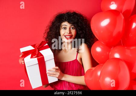 Feiertage und Feiern. Schöne Frau mit lockigen Haaren, in der Nähe von Herzballons stehen, Geschenkbox halten und glücklich lächeln, whi Stockfoto