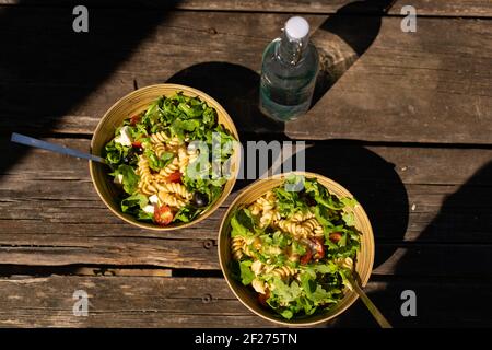 Griechischer mediterraner Salat mit Feta-Käse, Pasta, Oliven, Kirschtomaten und Rucola auf einer Bambusschüssel auf einem hölzernen Picknicktisch Stockfoto