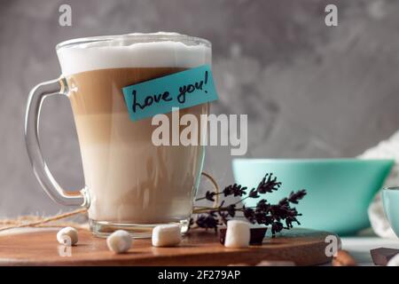 Kaffee Latte Tasse mit Love You Aufkleber auf Holztablett mit türkisfarbener Schale, auf grauem Wandhintergrund Stockfoto