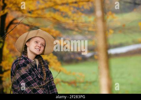 Eine junge Frau mit Strohhut und kariertem Hemd, lächelt, während sie draußen steht, mit verschwommenen Herbstbäumen hinter sich. Stockfoto