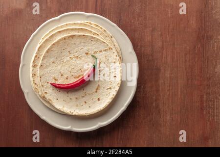 Ein roter, scharfer Chilischote, von oben auf einem Stapel von Tortillas, mexikanischen Fladenbroten, auf einem dunklen, rustikalen Holzhintergrund geschossen Stockfoto