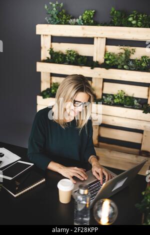 Hübsche, junge und blonde Frau mit stilvollen, modernen schwarzen Gläsern sitzt in einem nachhaltigen Büro und genießt einen Kaffee zu gehen Stockfoto