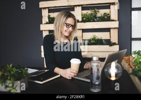 Hübsche, junge und blonde Frau mit stilvollen, modernen schwarzen Gläsern sitzt in einem nachhaltigen Büro und genießt einen Kaffee zu gehen Stockfoto