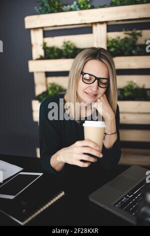 Hübsche, junge und blonde Frau mit stilvollen, modernen schwarzen Gläsern sitzt in einem nachhaltigen Büro und genießt einen Kaffee zu gehen Stockfoto