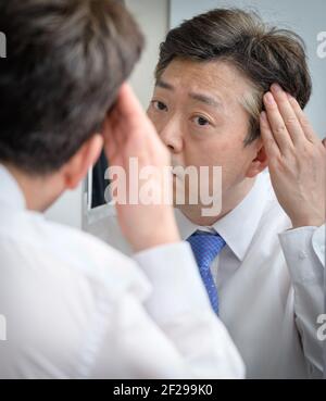 Asiatischer Mann mittleren Alters, der in den Spiegel schaut und seine berührt Haare Stockfoto