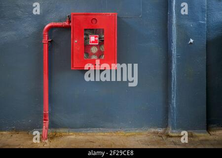 Feuerlöschschlauch in roter Box. Rohrrolle für den Brandfall in rotem Metallschrank auf grau lackierter Betonwand mit Fliesenboden als Teil der Brandbekämpfung Stockfoto